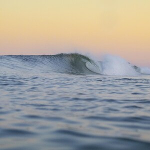 May include: A photograph of a breaking wave in the ocean at sunset. The wave is a translucent green colour, with white foam at the crest. The sky is a gradient of yellow and orange.