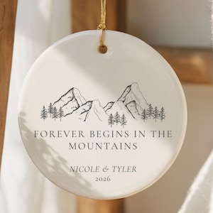 May include: A round white ceramic ornament hanging from a gold string. The front features a black line drawing of mountains and pine trees, with the text "FOREVER BEGINS IN THE MOUNTAINS", the names "NICOLE & TYLER", and the year "2026" printed below.