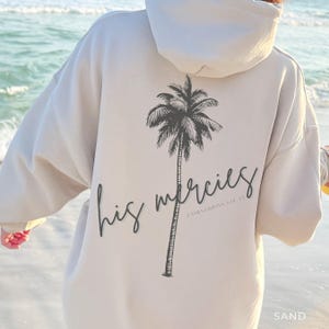 May include: A sand-colored hooded sweatshirt featuring a black graphic of a palm tree on the back. The text "his mercies" is written in a cursive script across the trunk, with "LAMENTATIONS 3:22-23" printed in smaller text below the script.