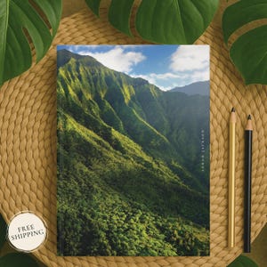 May include: A journal featuring a vibrant landscape image of a lush green mountain range under a blue sky with clouds. The journal has the text "SARAH JACKSON" printed vertically on the right side. Two pencils, one gold and one black, are placed next to the journal.