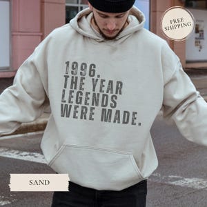 May include: Sand-colored hoodie with a front pocket and a drawstring hood. The front of the hoodie features the text "1996. THE YEAR LEGENDS WERE MADE." The hoodie is a neutral color and suitable for casual wear.
