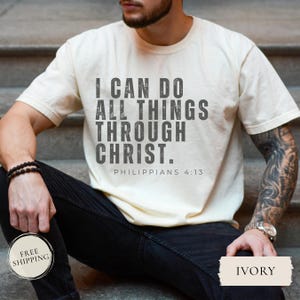 May include: Ivory-colored t-shirt with the text "I CAN DO ALL THINGS THROUGH CHRIST. Philippians 4:13" in gray. The shirt is short-sleeved and worn by a person sitting on steps. A watch and bracelet are visible.