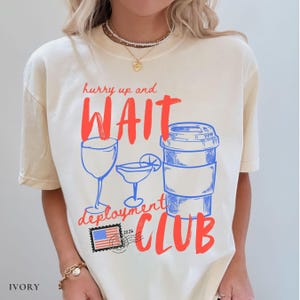 May include: Ivory-colored t-shirt with red and blue graphic text and illustrations. The text reads "hurry up and WAIT deployment CLUB" with images of a wine glass, cocktail, and coffee cup. A postage stamp with the American flag is also included.