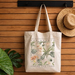 May include: A beige canvas tote bag featuring a botanical print of green tropical leaves and orange orchids. The text "LET EVERYTHING THAT HAS BREATH PRAISE THE LORD" and "PSALM 150:6" is printed in black capital letters above and below the floral design.