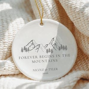May include: A round white ceramic ornament hanging from a gold string. The ornament features a black line drawing of mountains and pine trees. Below the illustration, the text reads FOREVER BEGINS IN THE MOUNTAINS, NICOLE & TYLER, 2026.