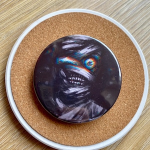 Könnte beinhalten: Runder Knopf mit einem dunklen, künstlerischen Design, das ein Wesen mit breitem, zahnigem Grinsen und einem einzelnen, farbenfrohen Auge zeigt. Der Knopf liegt auf einem Korkuntersetzer mit weißem Rand auf einer Holzoberfläche.