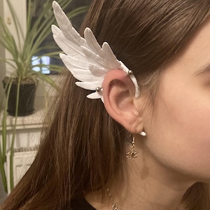 Op de afbeelding: Een witte veren oormanchet met een kleine gouden lotusbloem oorring. De oormanchet is bevestigd aan het oor met een zilveren stud.