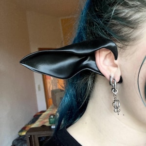 Könnte beinhalten: Eine schwarze, spitze Ohrprothese, die am Ohr getragen wird. Die Prothese ist glatt und glänzend, mit einer gebogenen Form. Die Person trägt ein schwarzes Oberteil und hat blaue Haare. Silberne Ohrringe sind sichtbar.