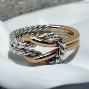 May include: A silver and gold intertwined knot ring. The ring is made of two bands, one silver and one gold, that are twisted together to form a knot.