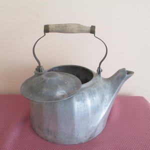 May include: A vintage metal kettle with a lid and spout. The kettle has a wooden handle and a curved metal handle. The kettle is silver and sits on a pink surface. The lid is partially open.