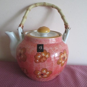 May include: A ceramic teapot with a bamboo handle and a floral design. The teapot is primarily pink with orange and white flower accents. The lid is yellow and white. The teapot has a white spout and a small label with Japanese characters.