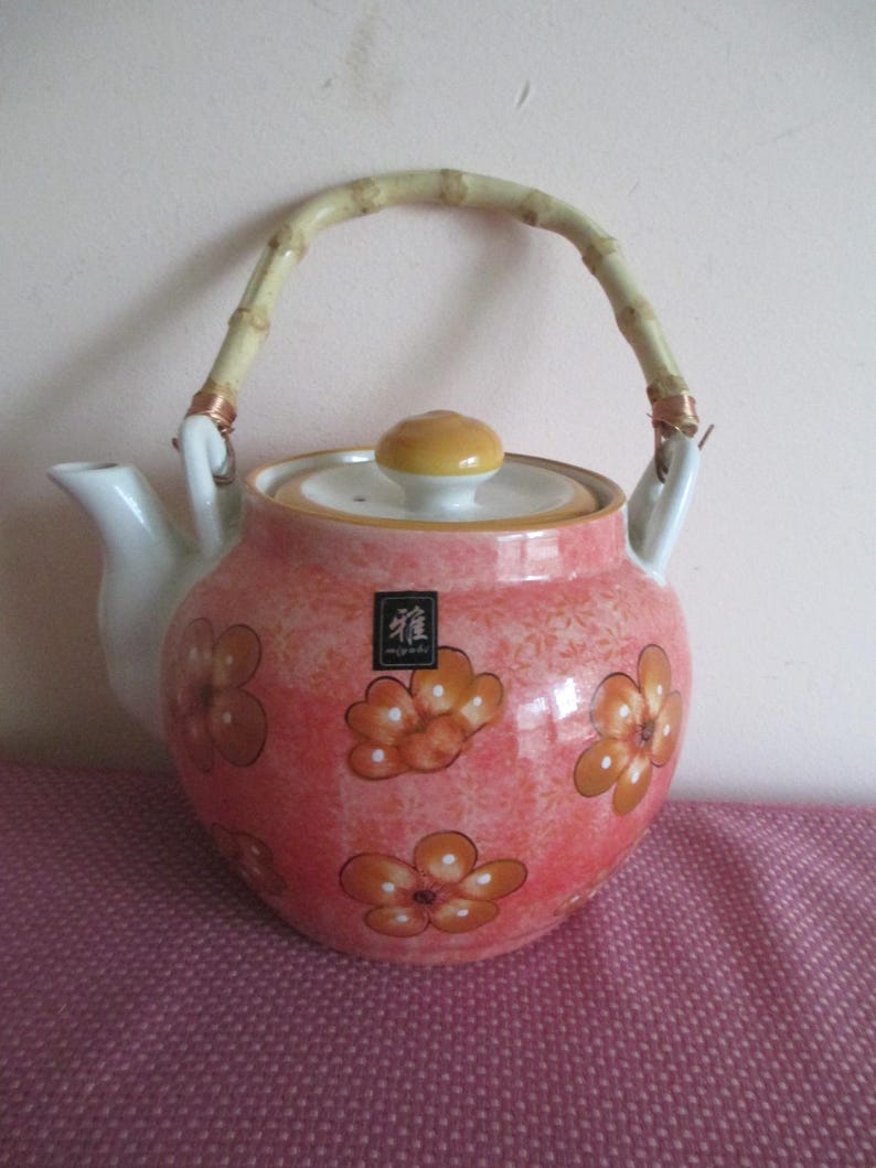 K&ouml;nnte beinhalten: Eine rosa Keramik-Teekanne mit Blumenmuster und Bambusgriff. Die Teekanne hat einen wei&szlig;en Ausguss und einen gelb-wei&szlig;en Deckel. Das Design umfasst orangefarbene und wei&szlig;e Blumen. Ein kleines Etikett mit japanischen Schriftzeichen ist sichtbar.