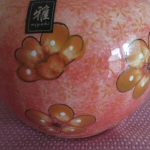 K&ouml;nnte beinhalten: Eine Keramikvase mit pfirsichfarbenem Hintergrund und orangefarbenen Blumendesigns. Die Vase hat eine gl&auml;nzende Oberfl&auml;che und ein kleines schwarzes Etikett mit japanischem Text. Die Blumen haben wei&szlig;e Tupfen.