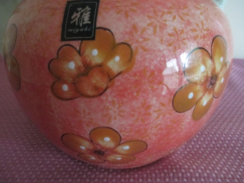 K&ouml;nnte beinhalten: Eine Keramikvase mit pfirsichfarbenem Hintergrund und orangefarbenen Blumendesigns. Die Vase hat eine gl&auml;nzende Oberfl&auml;che und ein kleines schwarzes Etikett mit japanischem Text. Die Blumen haben wei&szlig;e Tupfen.