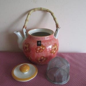K&ouml;nnte beinhalten: Rosa Keramik-Teekanne mit orangefarbenen Blumenmustern, Bambusgriff, Deckel und einem Tee-Sieb aus Metall. Ein dekoratives Tee-Set-Element. Ein charmanter Zusatz f&uuml;r jedes Teeservice.