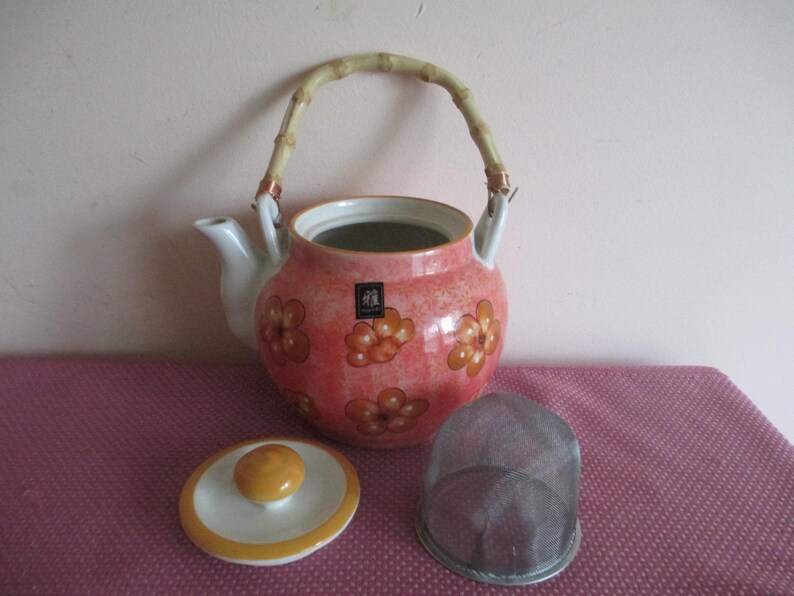 K&ouml;nnte beinhalten: Rosa Keramik-Teekanne mit orangefarbenen Blumenmustern, Bambusgriff, Deckel und einem Tee-Sieb aus Metall. Ein dekoratives Tee-Set-Element. Ein charmanter Zusatz f&uuml;r jedes Teeservice.