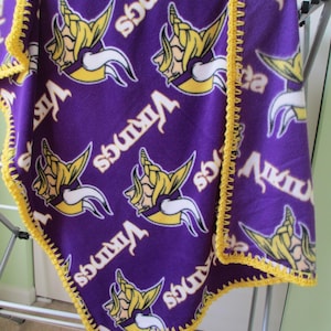 May include: A purple fleece blanket with the Minnesota Vikings logo in yellow, white, and purple. The blanket has a yellow crocheted border and the word "Vikings" printed on it. The blanket is draped over a clothes airer.