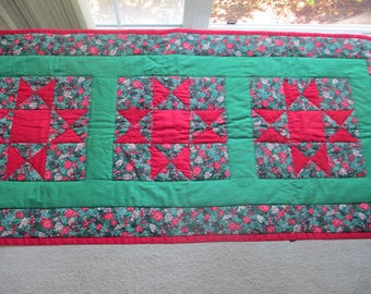 May include: A quilted table runner featuring a green background with three red star-shaped designs. The stars are surrounded by floral fabric. The edges are trimmed with a floral pattern and red border.