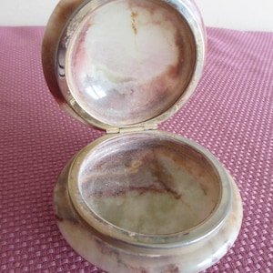 May include: Open, round jewelry box with a hinged lid. The box is made of a marbled stone with shades of cream, brown, and pink. A silver-colored metal band frames the top and bottom. The interior is a lighter color.