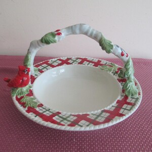 May include: A white ceramic serving dish with a handle, featuring a red cardinal bird and a decorative rim with red and green plaid patterns. The handle is adorned with green leaves and red berries.