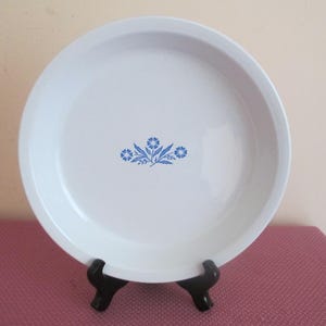 May include: White pie plate with a blue floral design in the center. The plate is round with a slightly raised edge and is displayed on a black stand. The floral design features blue flowers and leaves.