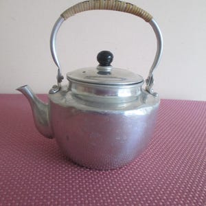 May include: A vintage silver-colored teapot with a curved handle wrapped in woven material. The teapot has a spout, a lid with a black knob, and sits on a pink surface. The teapot is made of metal and is likely used for brewing tea.