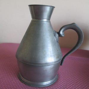 May include: A vintage pewter pitcher with a conical body, flared spout, and a curved handle. The pitcher has a dark, aged appearance, suggesting it is an antique. The handle is attached to the body with a decorative element.
