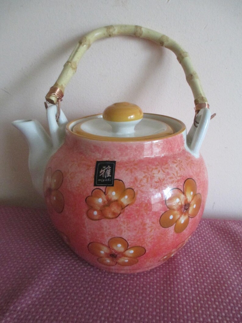K&ouml;nnte beinhalten: Rosa Keramik-Teekanne mit orangefarbenen Blumenmustern und einem Bambusgriff. Der Griff ist mit Kupferdraht befestigt. Der Deckel ist gelb und wei&szlig;. Zum Servieren von Tee.