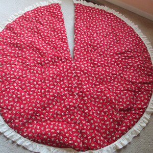 May include: Red quilted Christmas tree skirt with a white ruffled edge. The skirt is decorated with a small white floral pattern.