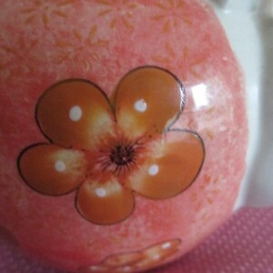 K&ouml;nnte beinhalten: Nahaufnahme eines Keramikobjekts mit rosa Hintergrund und einem gro&szlig;en orangefarbenen Blumendesign. Die Blume hat f&uuml;nf Bl&uuml;tenbl&auml;tter mit wei&szlig;en Punkten und einem dunkelbraunen Zentrum. Das Objekt hat einen wei&szlig;en Abschnitt.