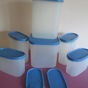 May include: A set of five translucent rectangular food storage containers with blue lids, including one tall container, two medium containers, and two smaller containers, plus two additional detached blue lids placed in the foreground.