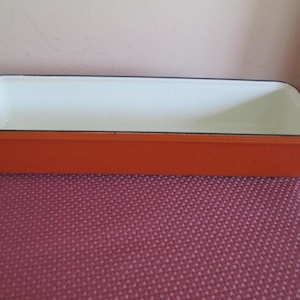 May include: A rectangular loaf pan with a vibrant orange exterior and a smooth white interior. The baking dish features a wide, flat rim and integrated handles on both ends, with a dark trim outlining the top edge of the vessel.