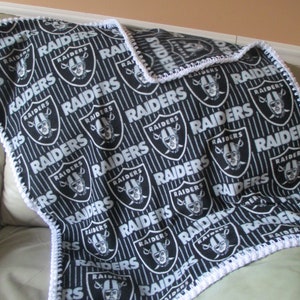 Peut inclure: Couverture Raiders noire et blanche avec un motif répété du logo et du nom de l'équipe. La couverture a une bordure crochetée blanche. Idéal pour les fans des Raiders.