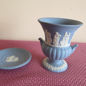 May include: A blue and white ceramic vase with a decorative design of figures and a small dog. The vase is on a pink and white patterned surface.
