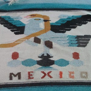 May include: A woven textile featuring a central graphic of an eagle with outstretched wings in shades of light blue, black, and tan. Below the eagle is a geometric design in red and blue, with the word MEXICO printed in red and black letters.