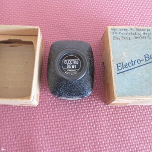 May include: An antique Electro-Bevi Standard device with its original packaging. The device is black with a circular dial. The packaging includes a box with text and handwritten notes. The items are on a pink surface.