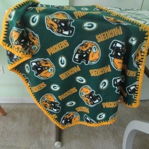 May include: Green and yellow fleece blanket with a crocheted border. The blanket features a repeated pattern of football helmets and the word "Packers".