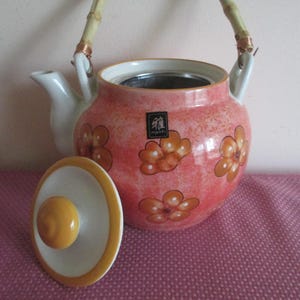K&ouml;nnte beinhalten: Eine lachsfarbene Keramik-Teekanne mit orangefarbenen Blumenmustern und einem Bambusgriff. Der Deckel ist separat. Die Teekanne hat einen wei&szlig;en Ausguss und Griffbefestigungen. Ma&szlig;e in Zentimetern.