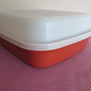 May include: A red and white plastic container with a lid. The container is rectangular with rounded corners. The lid is white and fits snugly on top. The container is sitting on a pink surface.