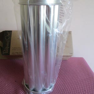 May include: A silver-colored metal scalloped bread mold with a cylindrical shape, enclosed in a clear plastic bag. Behind the mold is a cardboard box labeled The Pampered Chef, featuring the text Scalloped Bread Mold and Molde tubular.