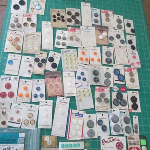 May include: A large collection of vintage buttons in various colors, sizes, and materials. The buttons are arranged on cards and in a jar. Some of the buttons have text on them, such as "Lansing" and "La Petite".