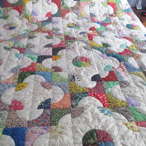 May include: A handmade quilt with a white background and colorful patchwork floral designs. The quilt features a repeating pattern of curved shapes and squares in various colors, including red, blue, green, and yellow. The quilt is likely made of cotton.