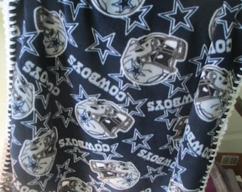 May include: A dark blue fleece blanket featuring a repeating pattern of white and silver football helmets, stars, and the text COWBOYS. The blanket has a white hand-stitched decorative edge finish along the entire perimeter of the fabric.