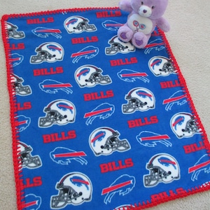 May include: Blue fleece baby blanket with a red crocheted border. The blanket features a repeating pattern of Buffalo Bills logos and the word "Bills". A purple teddy bear is sitting on the blanket.