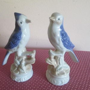 May include: Two ceramic bird figurines featuring white bodies with blue wings and tail feathers. Each bird is perched on a textured, light brown branch base with leaf details. The birds have small beaks and dark eyes, standing on circular white pedestals.