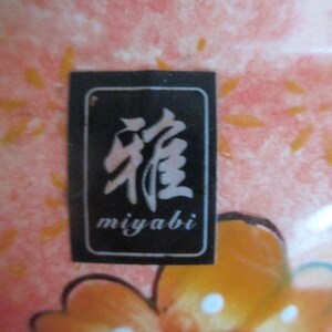 K&ouml;nnte beinhalten: Nahaufnahme eines Keramikst&uuml;cks mit einem rosa-orangenen Blumendesign. Ein schwarzes rechteckiges Etikett mit wei&szlig;en japanischen Schriftzeichen und dem Wort "miyabi" ist sichtbar.