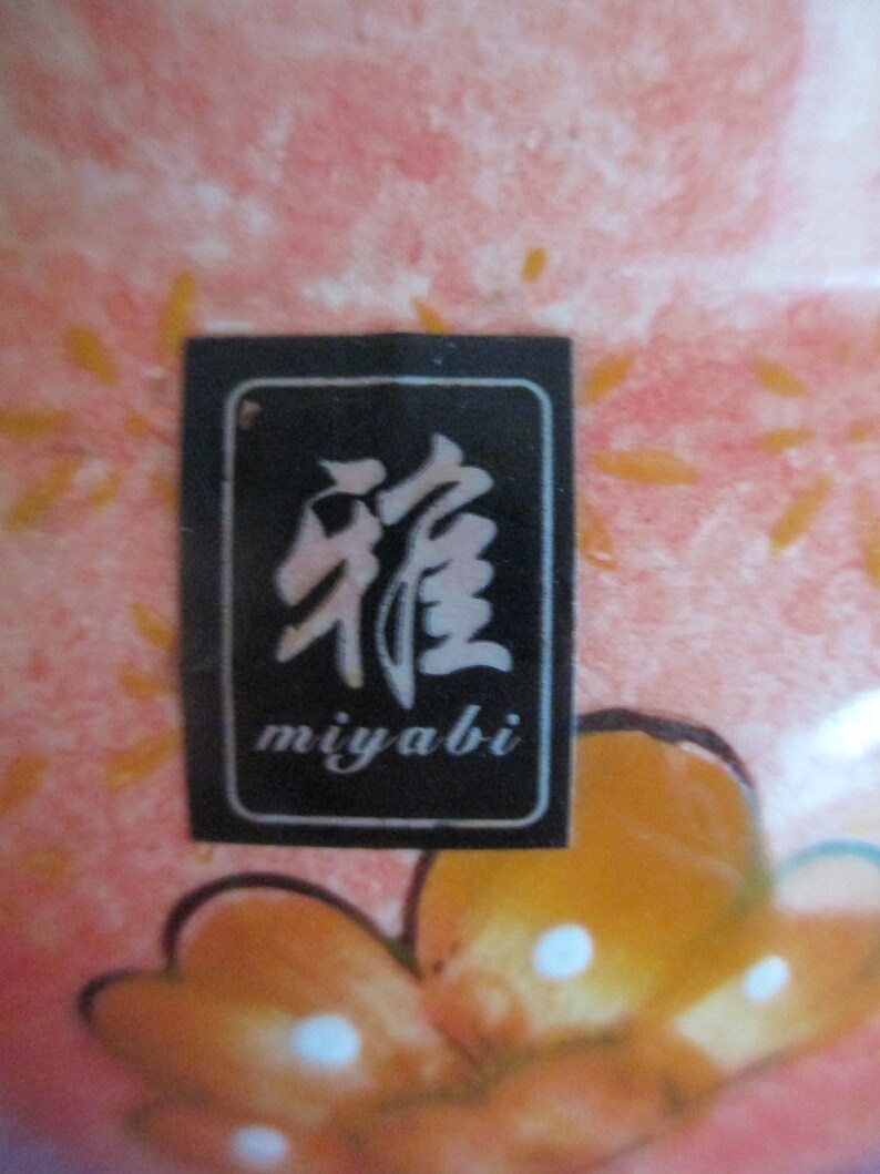 K&ouml;nnte beinhalten: Nahaufnahme eines Keramikst&uuml;cks mit einem rosa-orangenen Blumendesign. Ein schwarzes rechteckiges Etikett mit wei&szlig;en japanischen Schriftzeichen und dem Wort "miyabi" ist sichtbar.