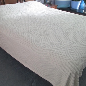 May include: A cream-colored bedspread featuring a raised, textured pattern of repeating circular and geometric shapes. The fabric has a consistent, soft-looking finish that covers the entire surface of the bed, draping over the sides.