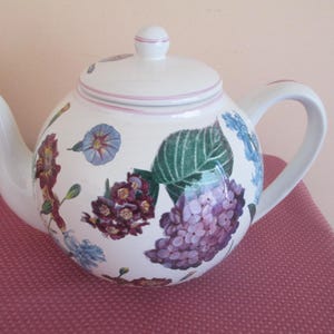 May include: White teapot with a floral design featuring pink, purple, blue, and green flowers. The teapot has a white lid with a pink trim.