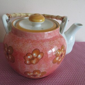 K&ouml;nnte beinhalten: Eine Keramik-Teekanne mit Bambusgriff und Deckel. Die Teekanne ist haupts&auml;chlich rosa mit einem Blumenmuster und orangefarbenen Blumendesigns. Der Ausguss und die Griffbefestigungen sind wei&szlig;.